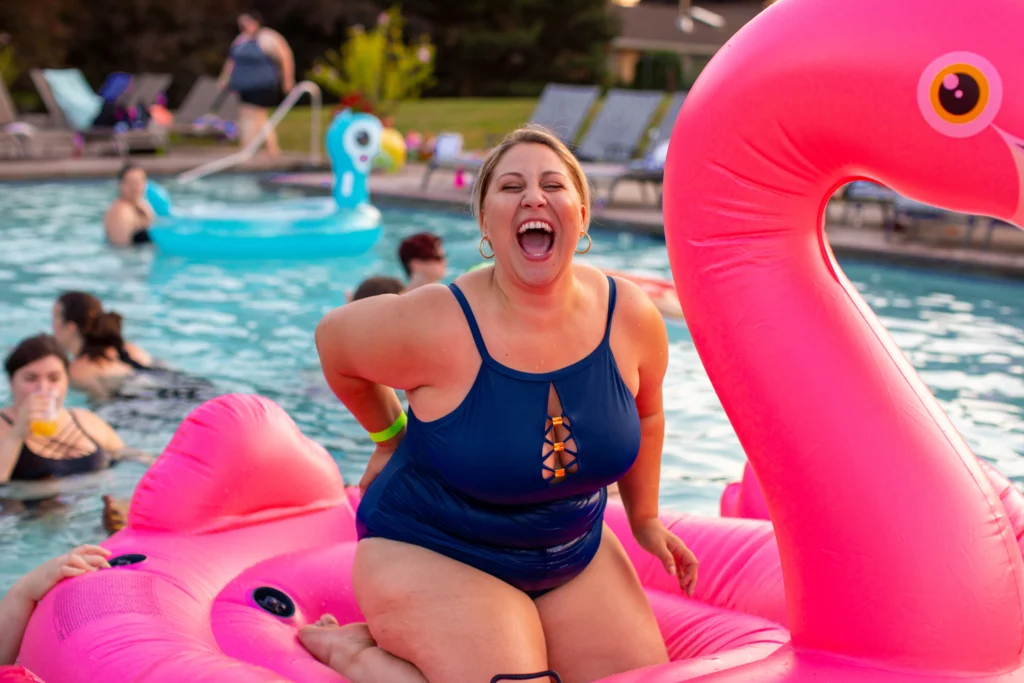 Dikke vrouw in een blauw badpak. Ze glimlacht breed en zit op een roze opblaasbare flamingo in het zwembad.